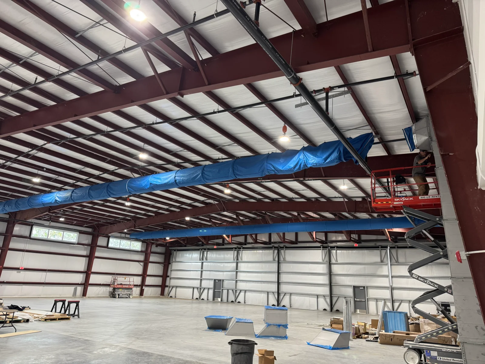 Blue insulated ductwork installed across steel beam structure in commercial warehouse with metal roof panels